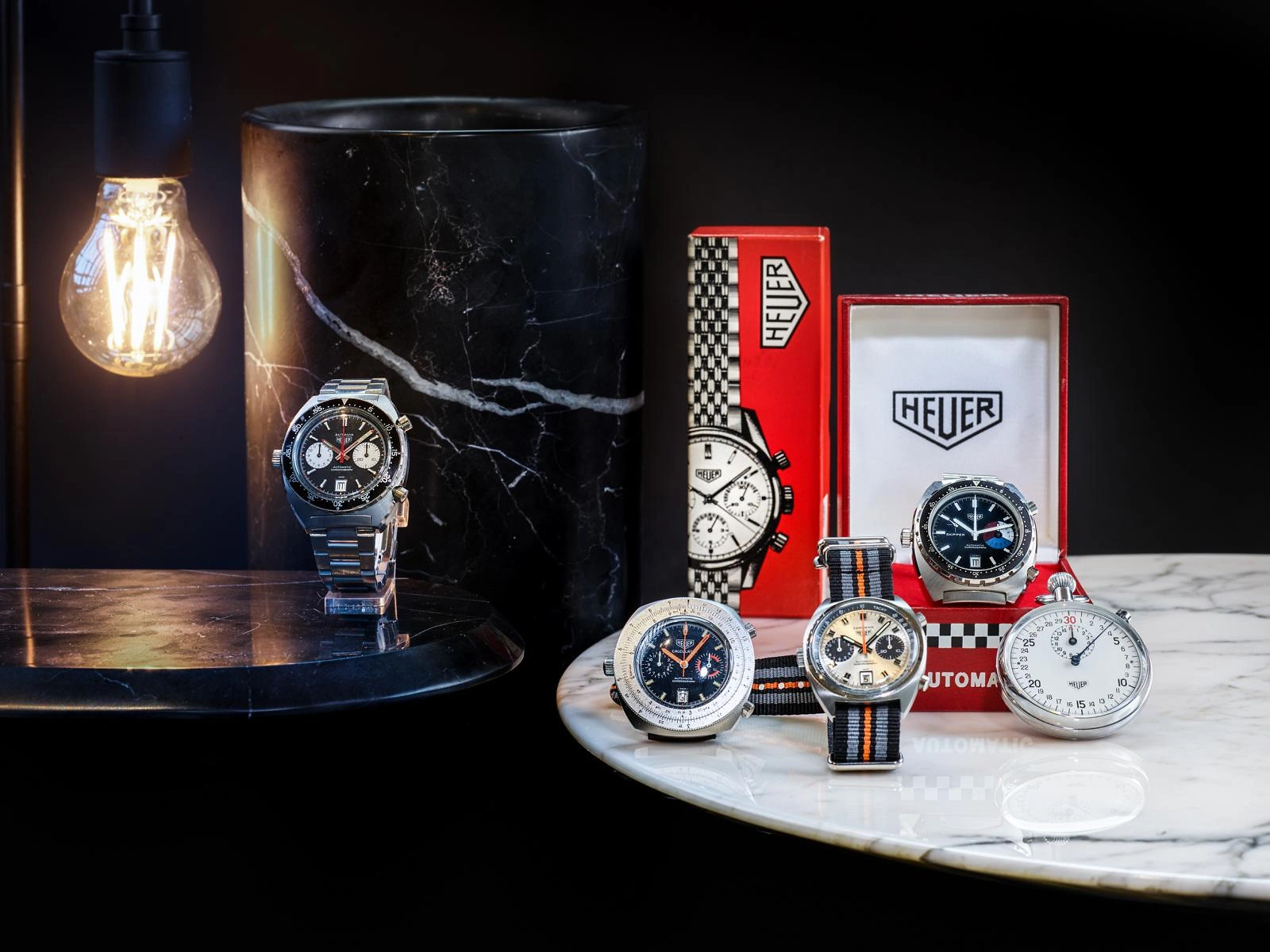 F&uuml;nf Tag Heuer Uhren sind stilvoll auf unterschiedlichen Marmorplatten arrangiert. Links im Bild steht eine Tischgl&uuml;hbirne, die warmes Licht auf die Szene wirft und die luxuri&ouml;se Pr&auml;sentation der Zeitmesser unterstreicht.